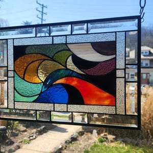 May include: Stained glass window panel with a black frame. The design features abstract shapes in shades of orange, red, blue, green, and brown. The panel is hanging from a chain and is displayed in front of a sunny outdoor scene.