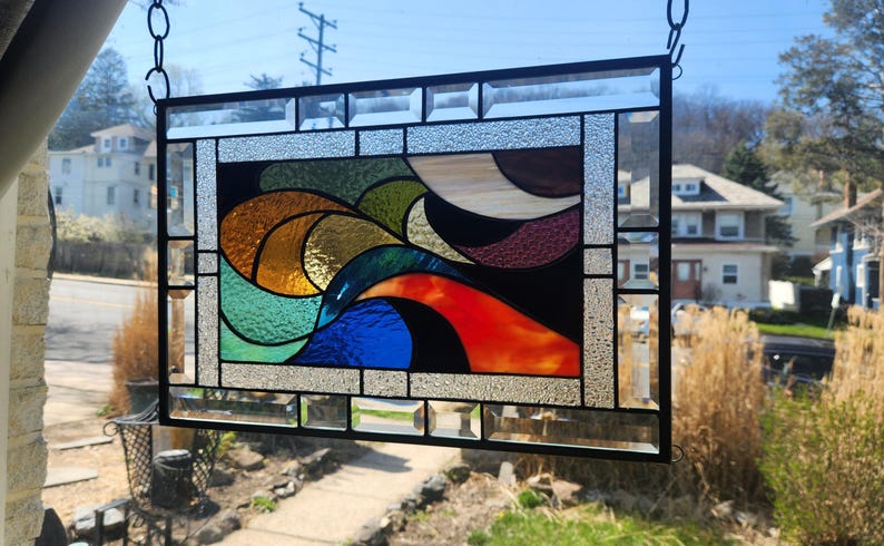 May include: Stained glass window panel with a black frame. The design features abstract shapes in shades of orange, red, blue, green, and brown. The panel is hanging from a chain and is displayed in front of a sunny outdoor scene.