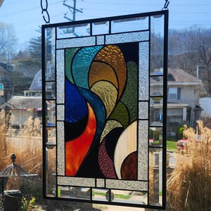 May include: Stained glass panel in a black metal frame, hanging from a chain. The abstract design uses vivid colours such as blue, orange, green, and gold. The glass has a textured finish, adding depth to the artwork.