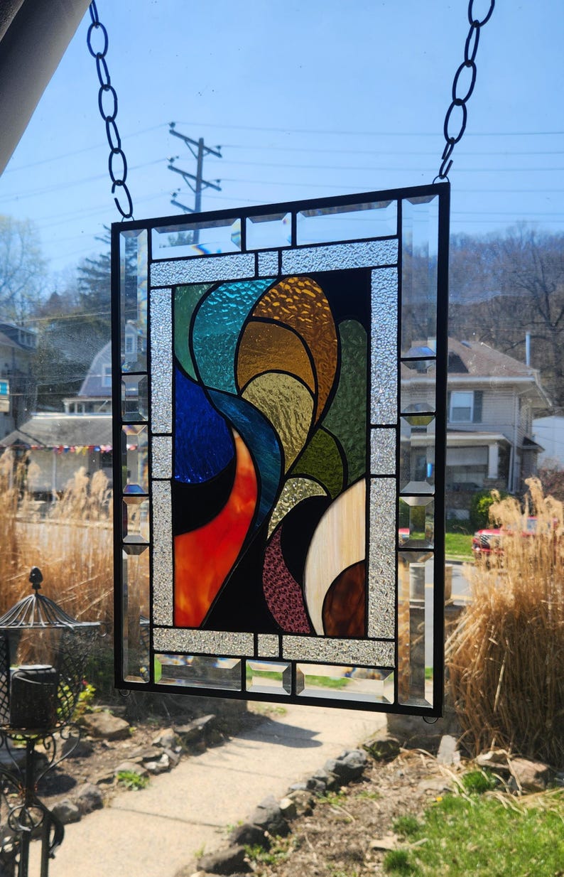 May include: Stained glass panel in a black metal frame, hanging from a chain. The abstract design uses vivid colours such as blue, orange, green, and gold. The glass has a textured finish, adding depth to the artwork.
