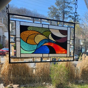 May include: A rectangular stained glass panel with a black metal frame, suspended by a chain. The panel features an abstract design with flowing shapes in shades of blue, orange, yellow, green, and brown. The glass has a textured appearance.