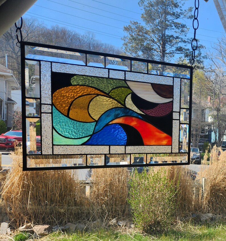 May include: A rectangular stained glass panel with a black metal frame, suspended by a chain. The panel features an abstract design with flowing shapes in shades of blue, orange, yellow, green, and brown. The glass has a textured appearance.