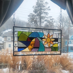 May include: A rectangular stained glass panel depicting a vibrant landscape with a large yellow flower and a stylized mountain range. The panel is framed in black and suspended by a chain, with a snowy outdoor scene visible through the window.