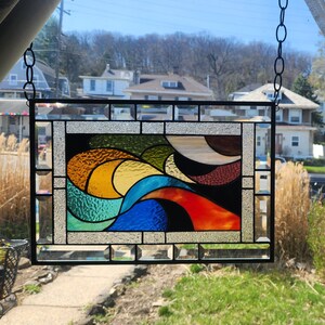 May include: Rectangular stained glass panel with a black metal frame, suspended by a chain. The design features an abstract composition of colourful glass pieces in shades of blue, orange, yellow, green, and brown.