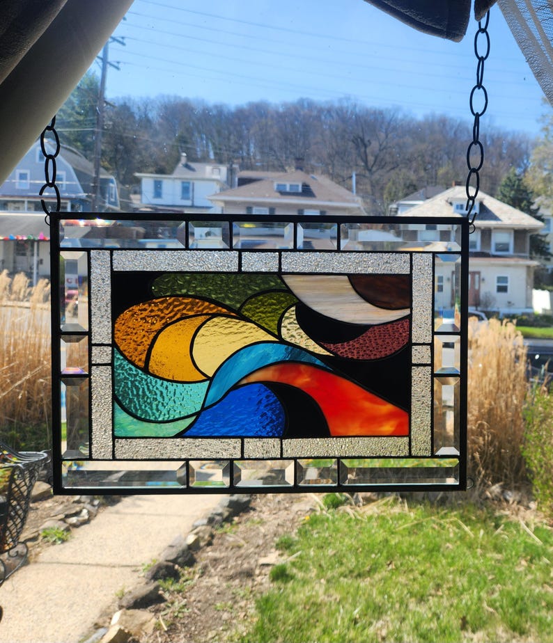 May include: Rectangular stained glass panel with a black metal frame, suspended by a chain. The design features an abstract composition of colourful glass pieces in shades of blue, orange, yellow, green, and brown.