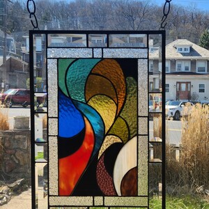 May include: A rectangular stained glass panel with a black metal frame, suspended by a chain. The design features abstract shapes in vibrant colours: red, blue, green, yellow, and brown. The glass has a textured appearance, and the panel is displayed outdoors.