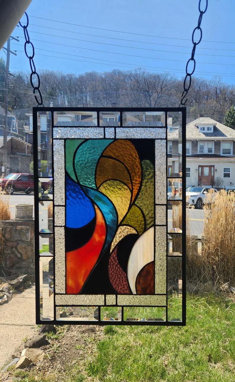 May include: A rectangular stained glass panel with a black metal frame, suspended by a chain. The design features abstract shapes in vibrant colours: red, blue, green, yellow, and brown. The glass has a textured appearance, and the panel is displayed outdoors.