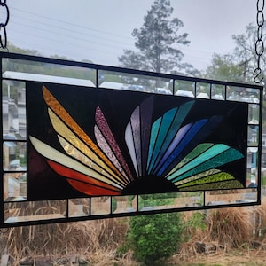 May include: A rectangular stained glass panel with a vibrant sunburst design. The sunburst features segments of orange, yellow, purple, blue, and green. The panel is framed in black and suspended by a chain, creating a decorative window art piece.