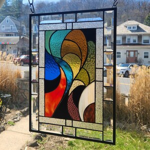 May include: A stained glass panel with a black metal frame, featuring an abstract design in blue, orange, yellow, green, and brown. The panel is suspended by a chain. The artwork is a vibrant and colourful piece, suitable for window display.