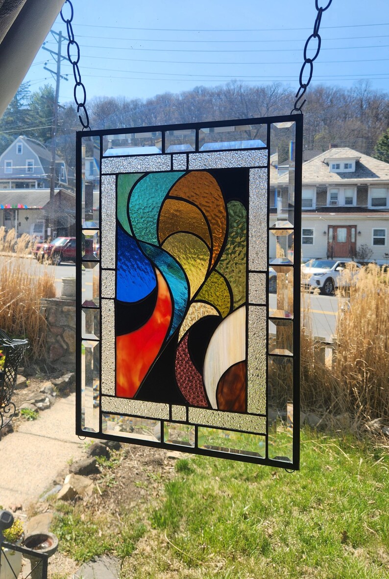 May include: A stained glass panel with a black metal frame, featuring an abstract design in blue, orange, yellow, green, and brown. The panel is suspended by a chain. The artwork is a vibrant and colourful piece, suitable for window display.