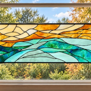 May include: A rectangular stained glass window hanging featuring a landscape design with rolling hills in shades of teal, turquoise, and blue, topped by a sky in warm tones of yellow, orange, and cream, all encased in a black metal frame.