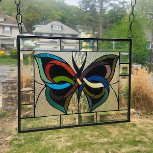 May include: Stained glass butterfly artwork in a black metal frame. The butterfly design incorporates vivid colours such as red, blue, green, and yellow. The artwork is suspended in front of a window, highlighting the intricate details.