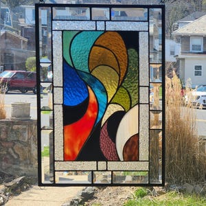 May include: Stained glass window panel with a black metal frame. The colourful design features abstract shapes in shades of red, orange, blue, green, and brown. The panel is hanging from a chain.