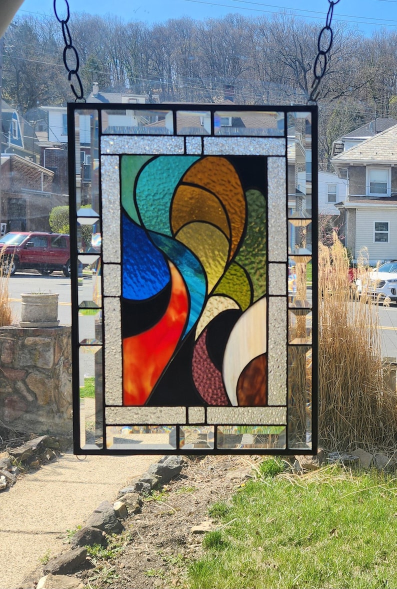 May include: Stained glass window panel with a black metal frame. The colourful design features abstract shapes in shades of red, orange, blue, green, and brown. The panel is hanging from a chain.
