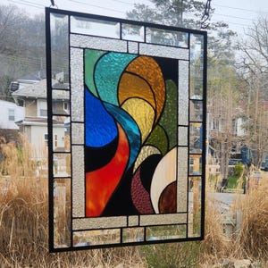 May include: A rectangular stained glass panel with a black metal frame. The design features abstract shapes in shades of blue, orange, yellow, green, and cream. The panel is suspended by a chain, showcasing the colourful artwork against a natural outdoor backdrop.