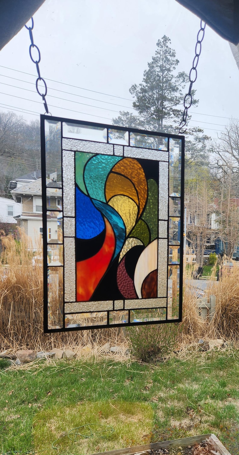 May include: A rectangular stained glass panel with a black metal frame. The design features abstract shapes in shades of blue, orange, yellow, green, and cream. The panel is suspended by a chain, showcasing the colourful artwork against a natural outdoor backdrop.