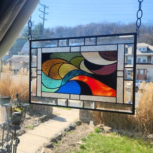 May include: A rectangular stained glass window with a black metal frame. The artwork features abstract shapes in shades of blue, green, orange, yellow, and brown. The window is hanging from a chain in front of a window with a view of houses and trees.