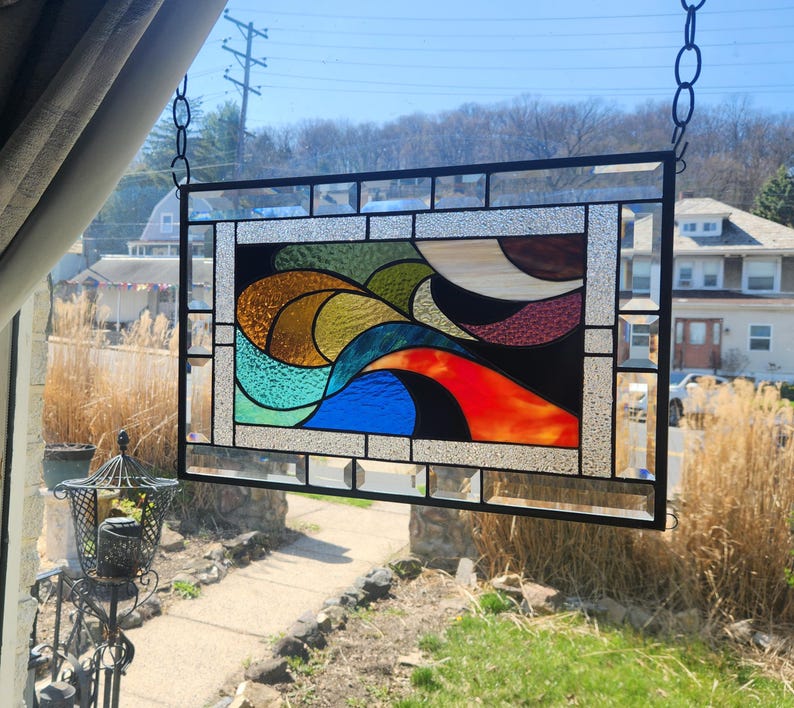 May include: A rectangular stained glass window with a black metal frame. The artwork features abstract shapes in shades of blue, green, orange, yellow, and brown. The window is hanging from a chain in front of a window with a view of houses and trees.