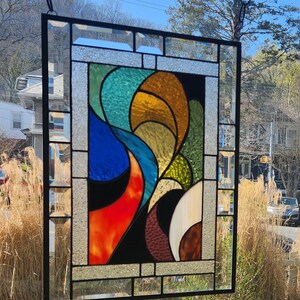 May include: A stained glass panel with a black frame and a colourful abstract design. The design features flowing shapes in shades of blue, orange, yellow, green, and brown. The panel is hanging outside, with a chain and the sky visible in the background.
