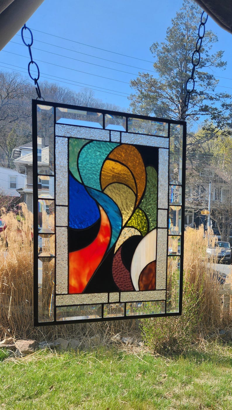May include: A stained glass panel with a black frame and a colourful abstract design. The design features flowing shapes in shades of blue, orange, yellow, green, and brown. The panel is hanging outside, with a chain and the sky visible in the background.
