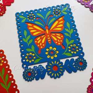 May include: A blue square paper cut-out with a butterfly design in orange and yellow, surrounded by green leaves and yellow flowers. The design is framed by a scalloped edge. Additional cut-outs with floral designs are in the background.