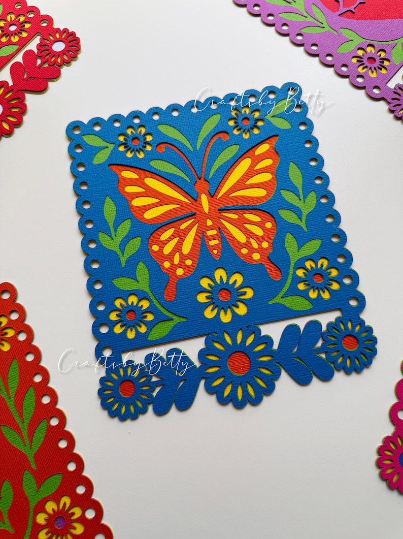 May include: A blue square paper cut-out with a butterfly design in orange and yellow, surrounded by green leaves and yellow flowers. The design is framed by a scalloped edge. Additional cut-outs with floral designs are in the background.