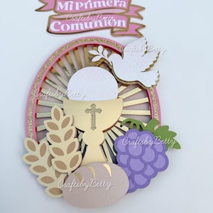 Op de afbeelding: Een gelaagde papieren taarttopper met een religieus thema. Het ontwerp omvat een gouden kelk met een kruis, een witte duif, een tros paarse druiven en de tekst "Mi Primera Comunión" op een roze banner. De achtergrond heeft een roze en gouden zonnestraalontwerp.