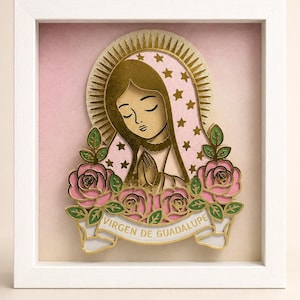 May include: A layered paper art piece of the Virgen de Guadalupe in a white frame. The figure has a gold halo, a pink mantle with gold stars, and hands in prayer. Below are pink roses with green leaves and a banner reading VIRGEN DE GUADALUPE.