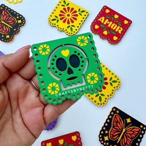 May include: A set of colorful paper cutouts with a skull design, a butterfly design, and the word "AMOR" in yellow letters on a red background. The cutouts are decorated with yellow and green flowers and hearts.