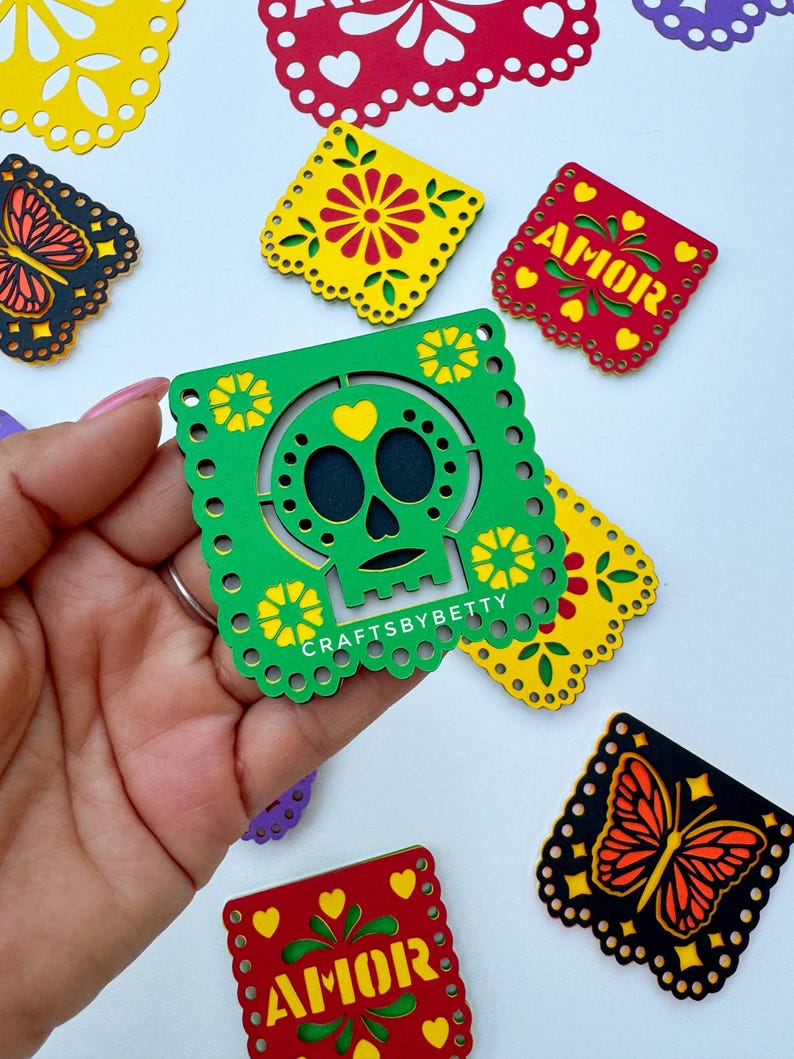 May include: A set of colorful paper cutouts with a skull design, a butterfly design, and the word "AMOR" in yellow letters on a red background. The cutouts are decorated with yellow and green flowers and hearts.
