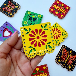 May include: A set of colorful paper cutouts with various designs, including butterflies, flowers, skulls, and the word "AMOR" in Spanish, which means "love". The cutouts are made of paper and have a scalloped edge. They are perfect for decorating for a party or celebration.