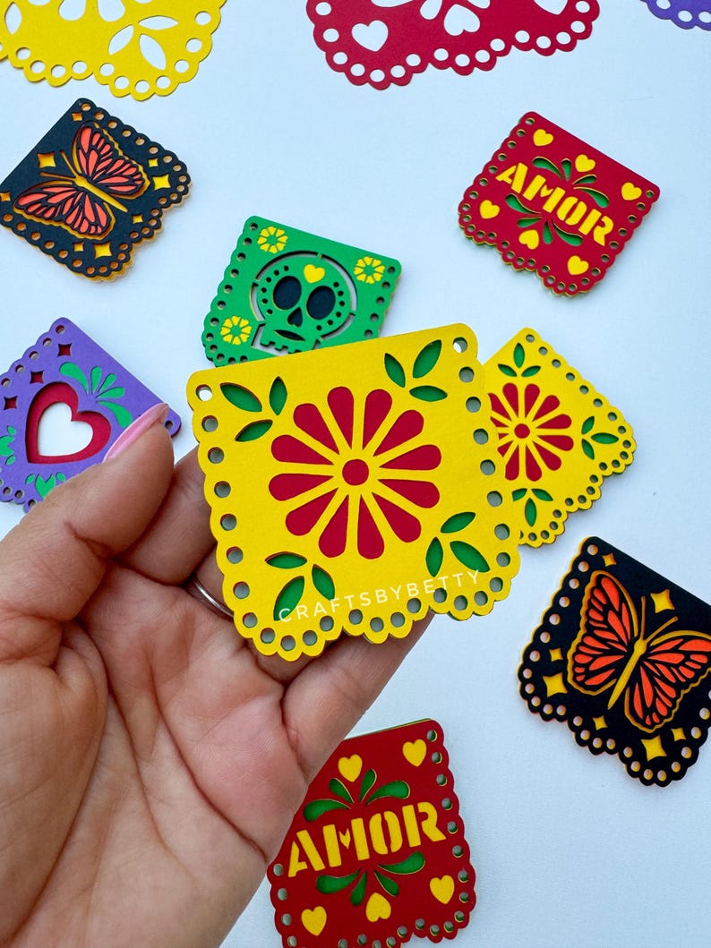 May include: A set of colorful paper cutouts with various designs, including butterflies, flowers, skulls, and the word "AMOR" in Spanish, which means "love". The cutouts are made of paper and have a scalloped edge. They are perfect for decorating for a party or celebration.