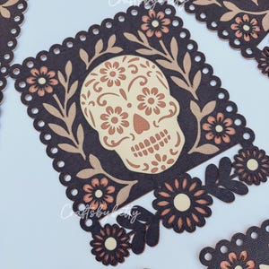 May include: Close-up of several Day of the Dead sugar skull cutouts. Each cutout features a skull design with floral accents in beige and brown tones, set against a dark brown background. The edges are scalloped.