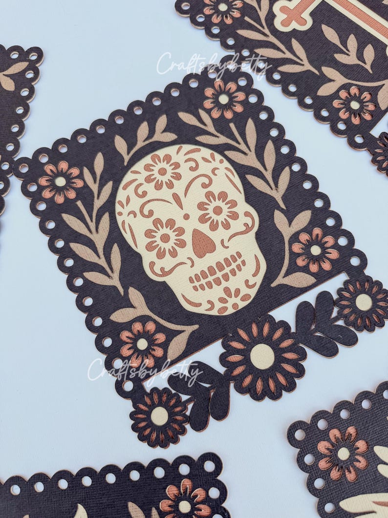 May include: Close-up of several Day of the Dead sugar skull cutouts. Each cutout features a skull design with floral accents in beige and brown tones, set against a dark brown background. The edges are scalloped.