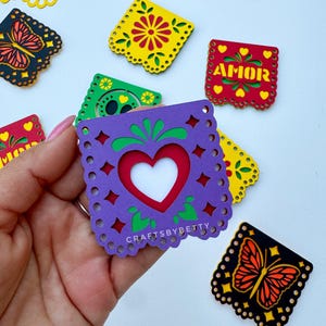 May include: A set of colorful paper cutouts with various designs, including butterflies, flowers, and hearts. The cutouts are decorated with yellow, red, green, purple, and black paper. The cutouts are labeled with the word "AMOR" in Spanish, which means "love".