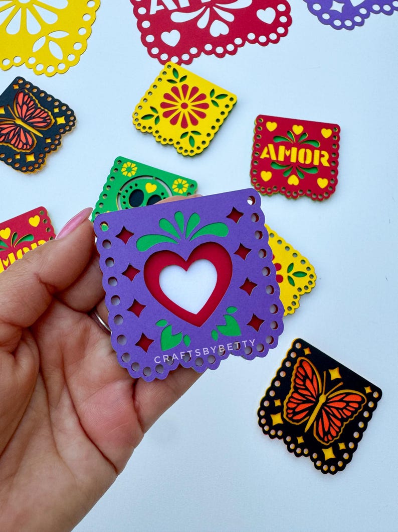 May include: A set of colorful paper cutouts with various designs, including butterflies, flowers, and hearts. The cutouts are decorated with yellow, red, green, purple, and black paper. The cutouts are labeled with the word "AMOR" in Spanish, which means "love".