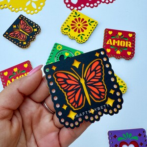 May include: Black paper cutout of a butterfly with orange and yellow wings. The cutout is decorated with a white dotted border and has the text "CRAFTSBYBETTY" in white letters. The cutout is part of a set of paper cutouts with various designs, including flowers and hearts.