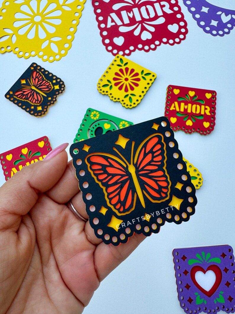 May include: Black paper cutout of a butterfly with orange and yellow wings. The cutout is decorated with a white dotted border and has the text "CRAFTSBYBETTY" in white letters. The cutout is part of a set of paper cutouts with various designs, including flowers and hearts.