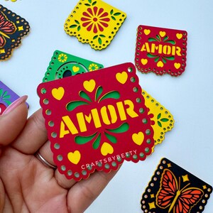 May include: A set of colorful paper cutouts with a butterfly, flowers, and the word "AMOR" in yellow letters on a red background. The cutouts are decorated with hearts and a scalloped edge.