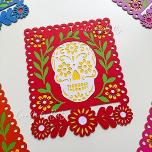 May include: A red paper cutout featuring a white sugar skull design with yellow flower accents, surrounded by green leaves and yellow flowers. The cutout is framed by a scalloped edge. Other colorful floral cutouts are in the background.