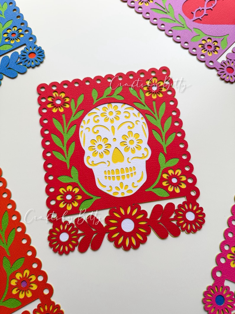 May include: A red paper cutout featuring a white sugar skull design with yellow flower accents, surrounded by green leaves and yellow flowers. The cutout is framed by a scalloped edge. Other colorful floral cutouts are in the background.