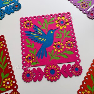 May include: A vibrant, layered paper art piece featuring a blue hummingbird on a pink background, surrounded by green leaves and yellow-centered flowers. The design is framed with a scalloped edge. The text "Crafted by Betty" is visible.