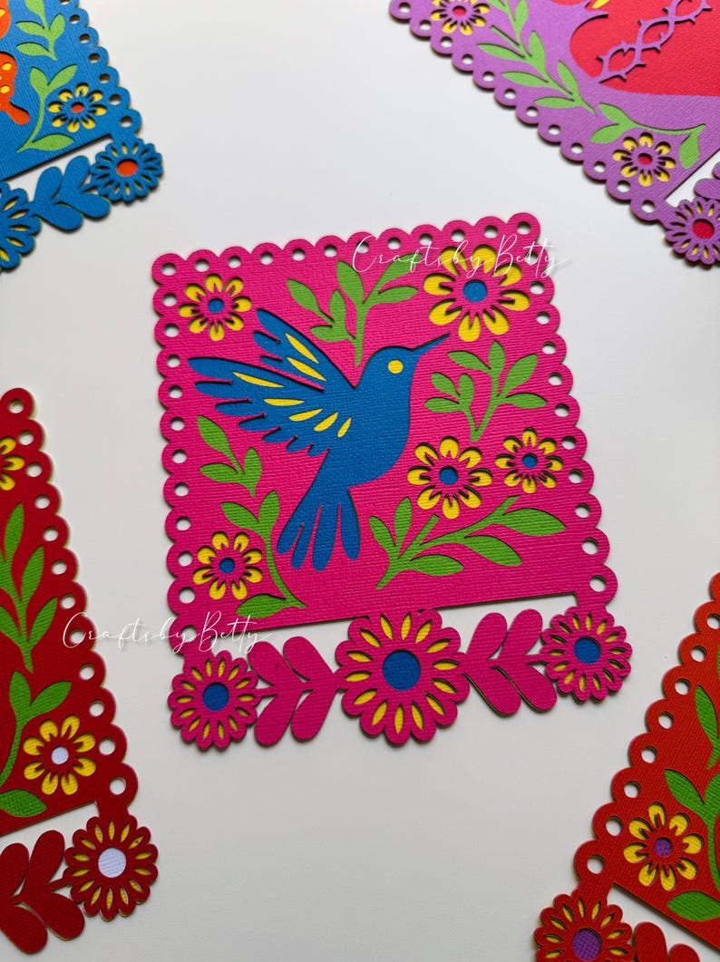 May include: A vibrant, layered paper art piece featuring a blue hummingbird on a pink background, surrounded by green leaves and yellow-centered flowers. The design is framed with a scalloped edge. The text "Crafted by Betty" is visible.