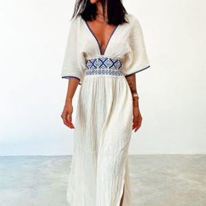 May include: A white maxi dress with a blue and white embroidered belt. The dress has a V-neckline and short sleeves.
