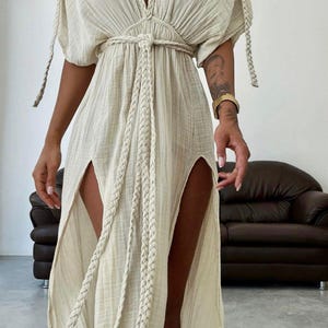 May include: A cream maxi dress with a deep V-neck and short sleeves. The dress has a braided belt and side slits. The sleeves and neckline are detailed with braided accents. The dress is made of a lightweight fabric.