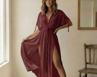 May include: A burgundy maxi dress with a deep V-neckline and high side slits. The dress features short sleeves with tie details and a braided waist tie. The model is standing in front of a window and a wooden chair.