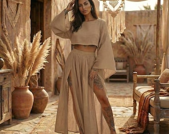 May include: A beige two-piece outfit featuring a cropped top with wide sleeves and a high-waisted skirt with side slits. The outfit is made of a lightweight fabric. The model is standing in a room with a bohemian aesthetic.