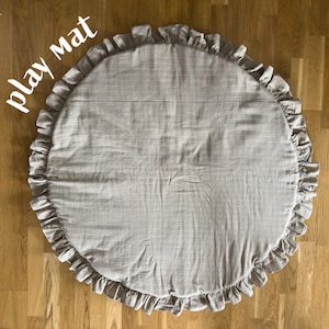 Könnte beinhalten: Ein runder, hellgrauer Spielteppich mit gerüschtem Rand. Der Teppich besteht aus einem weichen Stoff und liegt auf einem Holzboden. Die Worte "play mat" sind in weißer Schrift über dem Teppich geschrieben.