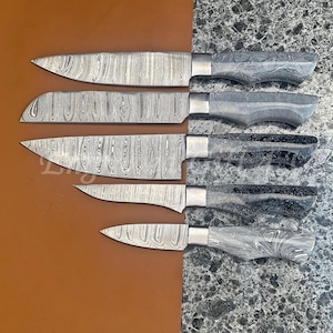 May include: A set of five Damascus steel knives with patterned blades and marbled gray handles. The blades have a distinctive wood grain appearance. The knives are arranged on a brown surface next to a gray speckled surface.