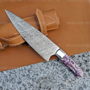 May include: A Damascus steel chef's knife with a purple and white handle. The knife is on a brown leather surface with a matching leather sheath.